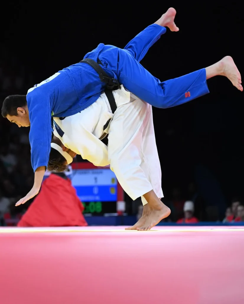 Een judoka in een wit judopak voert een Ippon Seoi Nage uit, waarbij de tegenstander in een blauw judopak over zijn schouder wordt geworpen tijdens een wedstrijd op een rood gevechtsoppervlak.