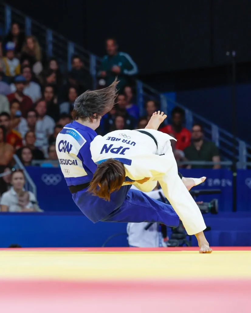 Een judoka voert een O-soto-gari worp uit tijdens een wedstrijd, waarbij de tegenstander gecontroleerd naar de grond wordt gebracht.