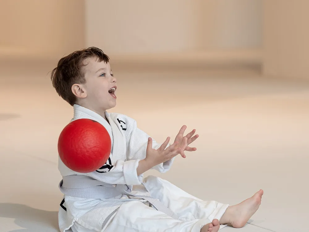 Tuimel Judo 3 Lachende kleuter in judopak vangt een rode bal tijdens Tuimel Judo bij Sportschool Herbert in Ede